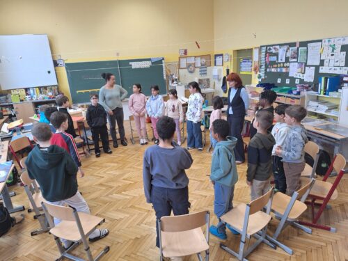 Kinder stehen im Kreis in der Klasse und hören der Vortragenden zu