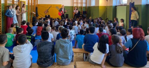 Kinder sitzen im Turnsaal und hören einem Mann und einer Frau beim musizieren zu